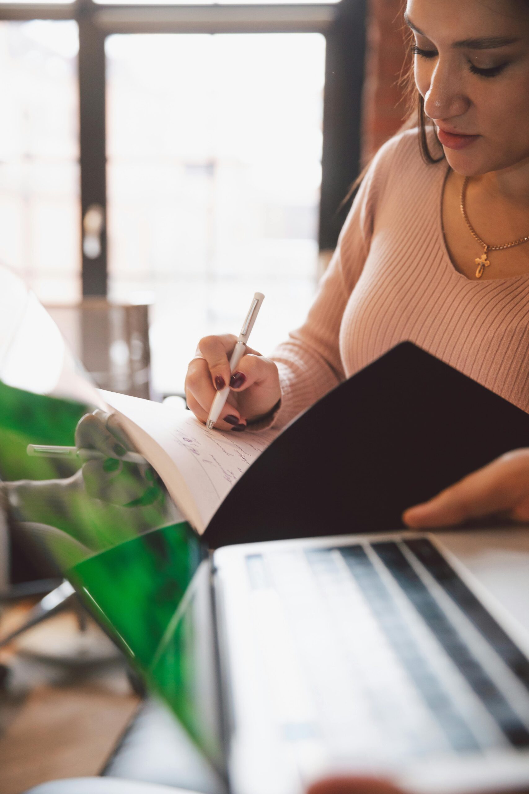 Woman holding a pen and writing in a notebook while sitting at a desk with a laptop.